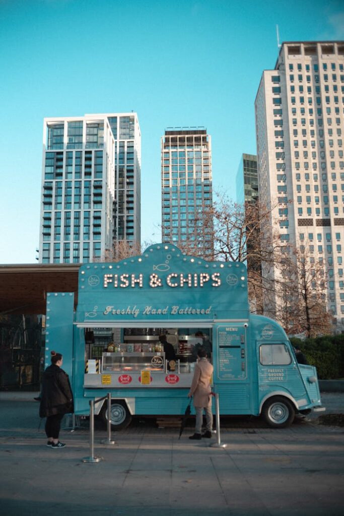 A vibrant blue food truck serves fish & chips amid urban skyscrapers, capturing urban life.